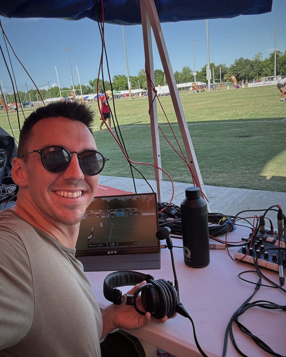 Andrew Rusk smiling at the commentary desk at Tropical 7s in Tampa, with headphones, a mixer and microphones beside the rugby field